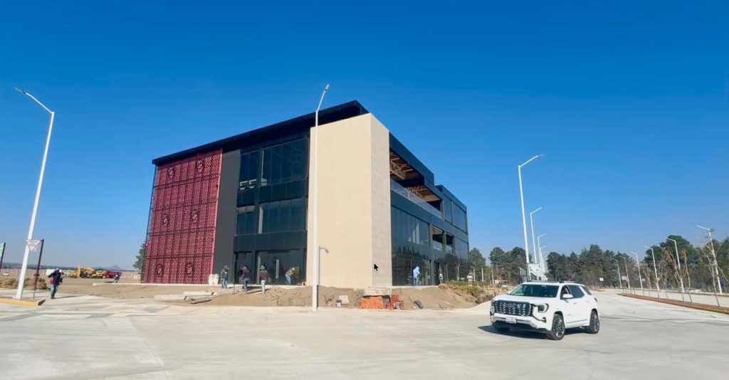 Edificio moderno de varios pisos con fachada de vidrio y estructura de color oscuro, situado en un área despejada con un automóvil blanco estacionado en primer plano.