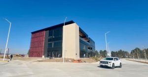 Edificio moderno de varios pisos con fachada de vidrio y estructura de color oscuro, situado en un área despejada con un automóvil blanco estacionado en primer plano.