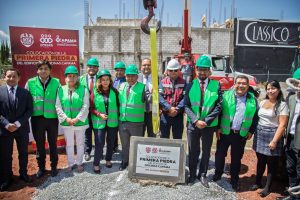 Tage de alt: Grupo de personas en un sitio de construcción durante una ceremonia de colocación de la primera piedra en Tlaxcala, México, con señalización y maquinaria en el fondo.