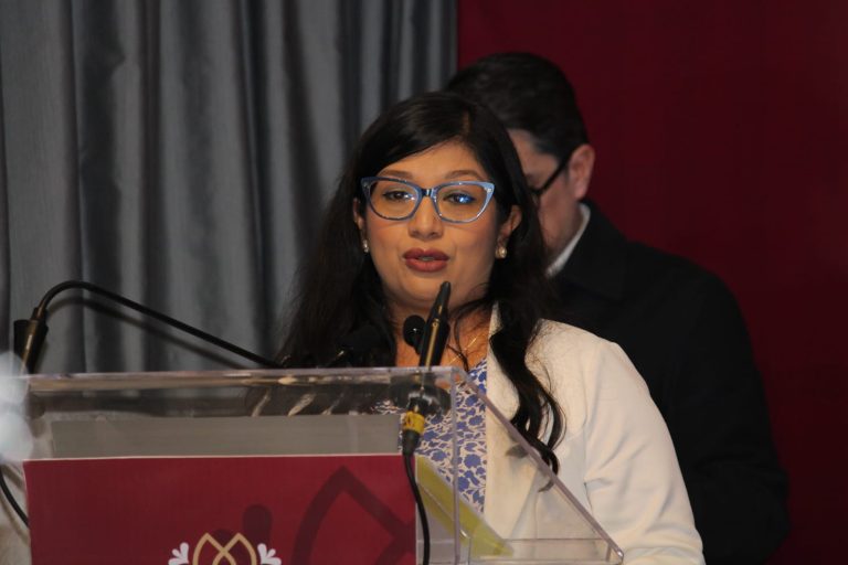 Mujer hablando en un evento formal, usando gafas y vestido tradicional, con fondo de cortinas grises y otra persona en la sombra, en un entorno de conferencia o presentación en Tlaxcala.