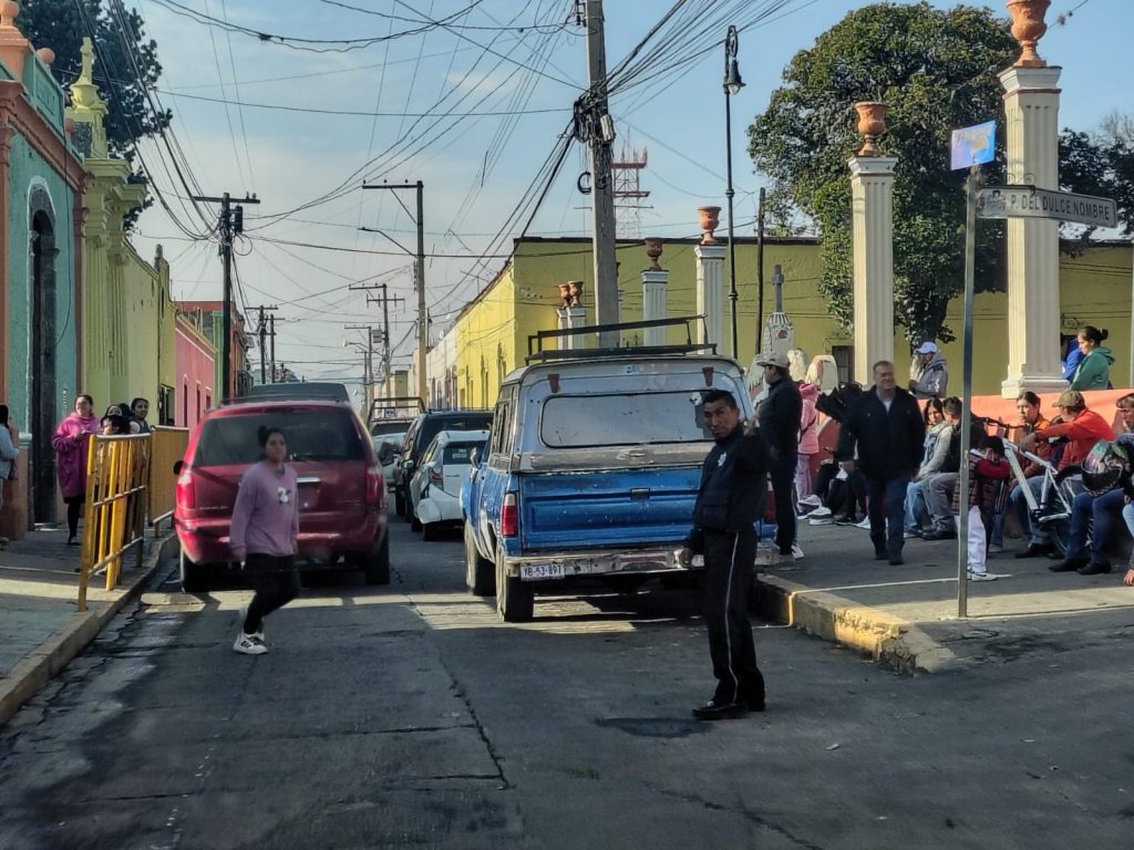 Calle en Tlaxcala con vehículos estacionados y personas esperando, en un día soleado.
