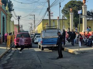 Calle en Tlaxcala con vehículos estacionados y personas esperando, en un día soleado.