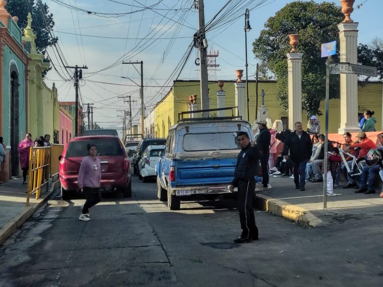 Calle en Tlaxcala con vehículos estacionados y personas esperando, en un día soleado.