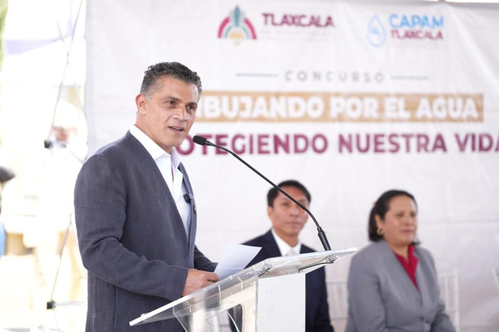 Hombre hablando en un podio durante un evento sobre agua y conservación en Tlaxcala, con un banner de fondo que promueve el cuidado del agua y la vida.