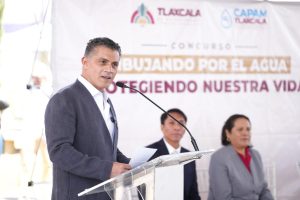 Hombre hablando en un podio durante un evento sobre agua y conservación en Tlaxcala, con un banner de fondo que promueve el cuidado del agua y la vida.