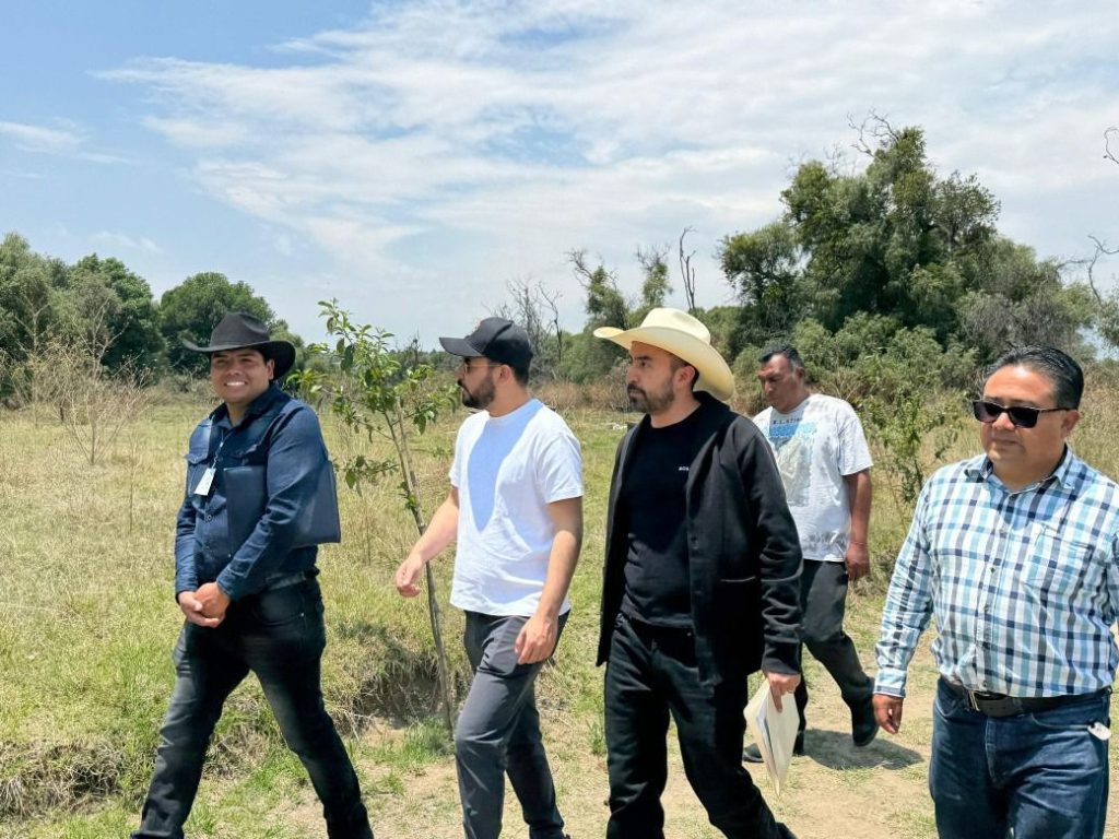Grupo de personas caminando en campo abierto con árboles y cielo despejado, en un día soleado.