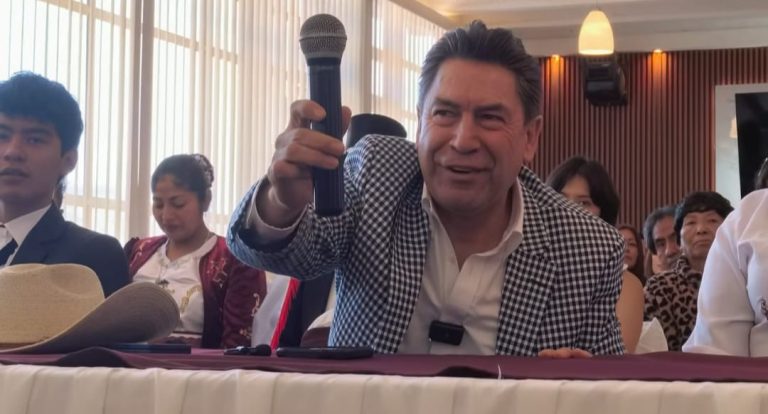 Hombre hablando en un evento, sosteniendo un micrófono, rodeado de varias personas en un ambiente interior con cortinas y luz cálida.