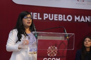 Mujer hablando en un evento político con fondo rojo y cartel que dice "EL PUEBLO MAN".