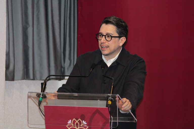 Persona hablando en un podio durante un evento formal, con fondo de cortinas grises y rojo.