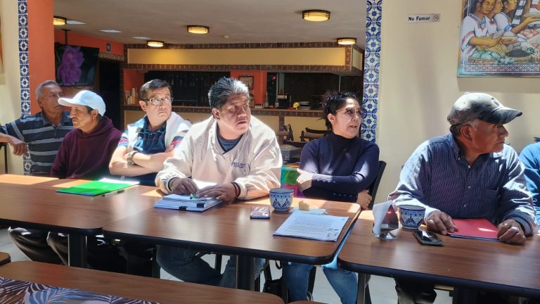 Reunión de adultos mayores en un espacio interior, participando en una actividad grupal, sentados en una mesa con documentos y tazas, en un ambiente decorado con arte y detalles culturales.
