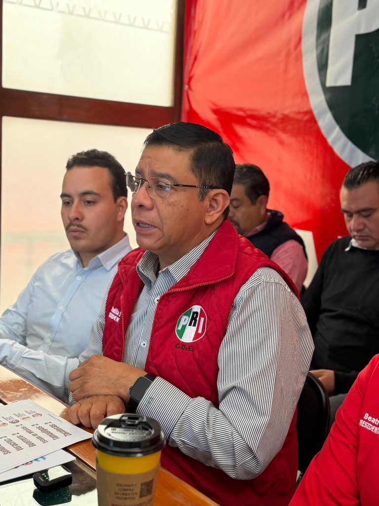 Hombre en reunión política con logotipo del PRI en fondo rojo, participando en un evento oficial en interiores.