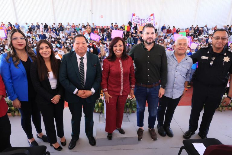 Grupo de personas en un evento público con una multitud de asistentes y pancartas en el fondo, en un espacio cerrado y bien iluminado.