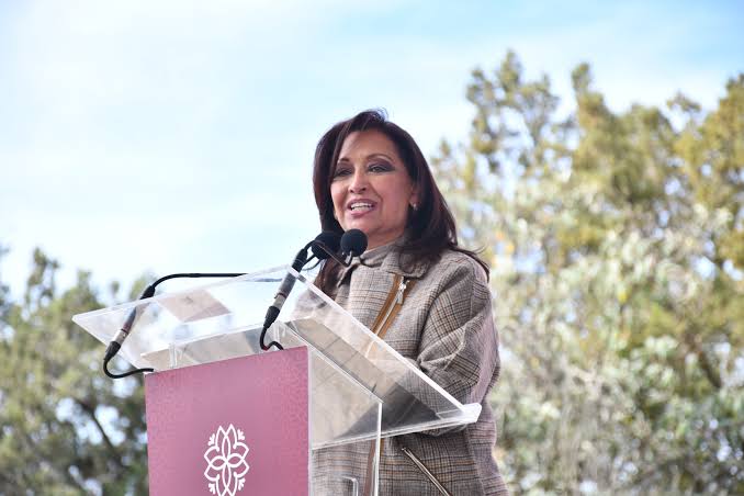 Mujer hablando en un podio al aire libre durante un evento público, con árboles y cielo en el fondo.