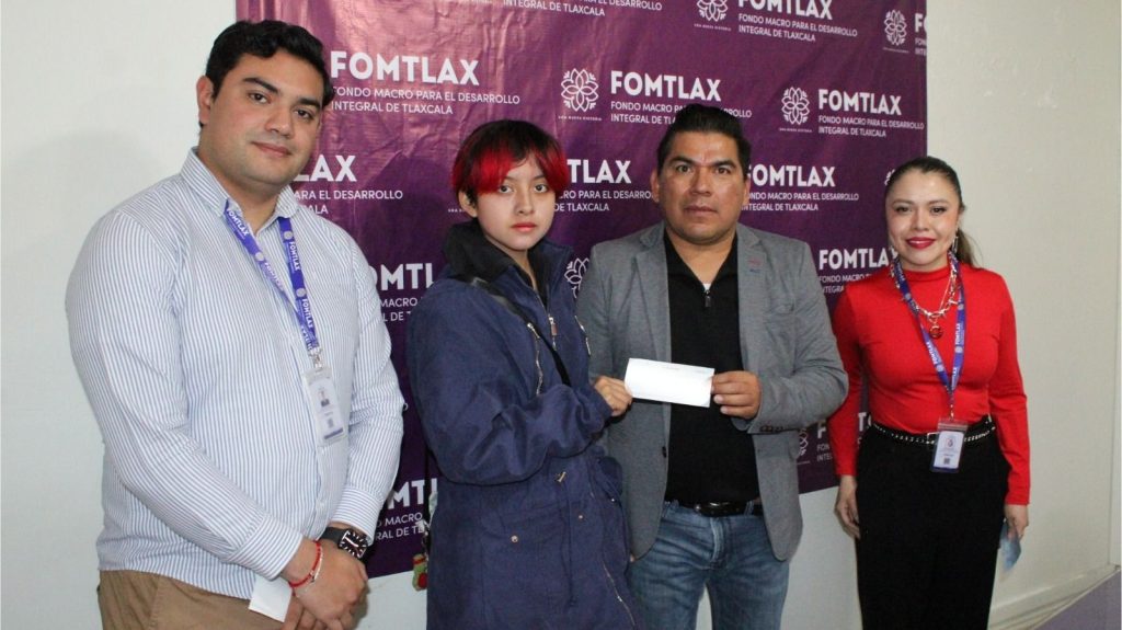 Persona entregando un cheque en un evento de FOMTLAX, con fondo con logotipos y texto relacionados con el desarrollo de Tlaxcala.