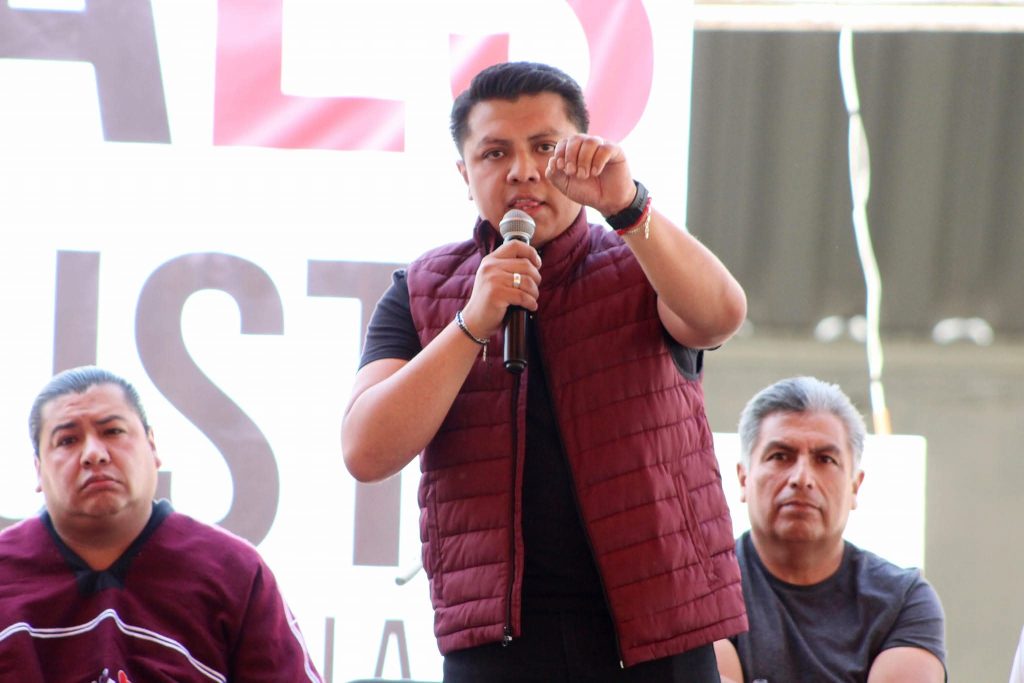 Mujer hablando en un micrófono durante un evento público con dos hombres sentados a su lado, en un espacio abierto con estructura metálica y cartel de fondo.
