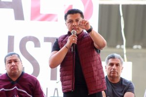 Mujer hablando en un micrófono durante un evento público con dos hombres sentados a su lado, en un espacio abierto con estructura metálica y cartel de fondo.