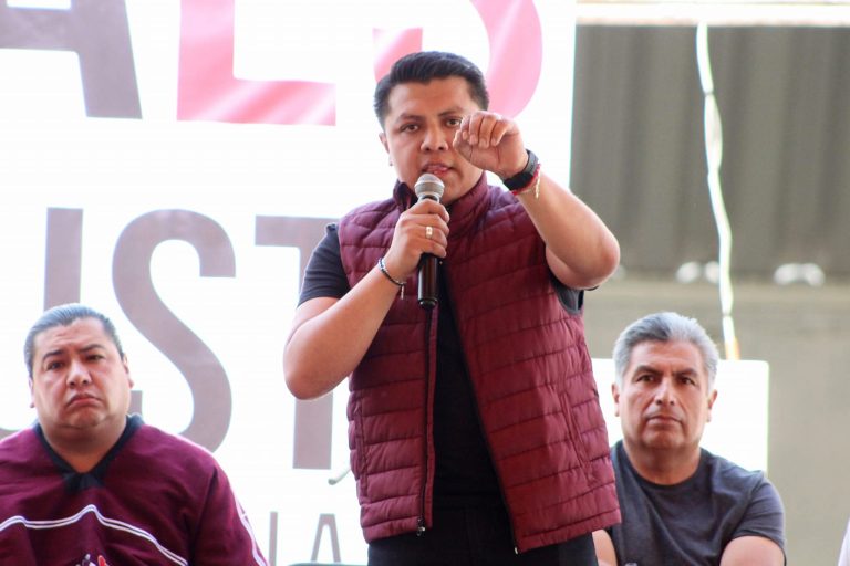 Mujer hablando en un micrófono durante un evento público con dos hombres sentados a su lado, en un espacio abierto con estructura metálica y cartel de fondo.