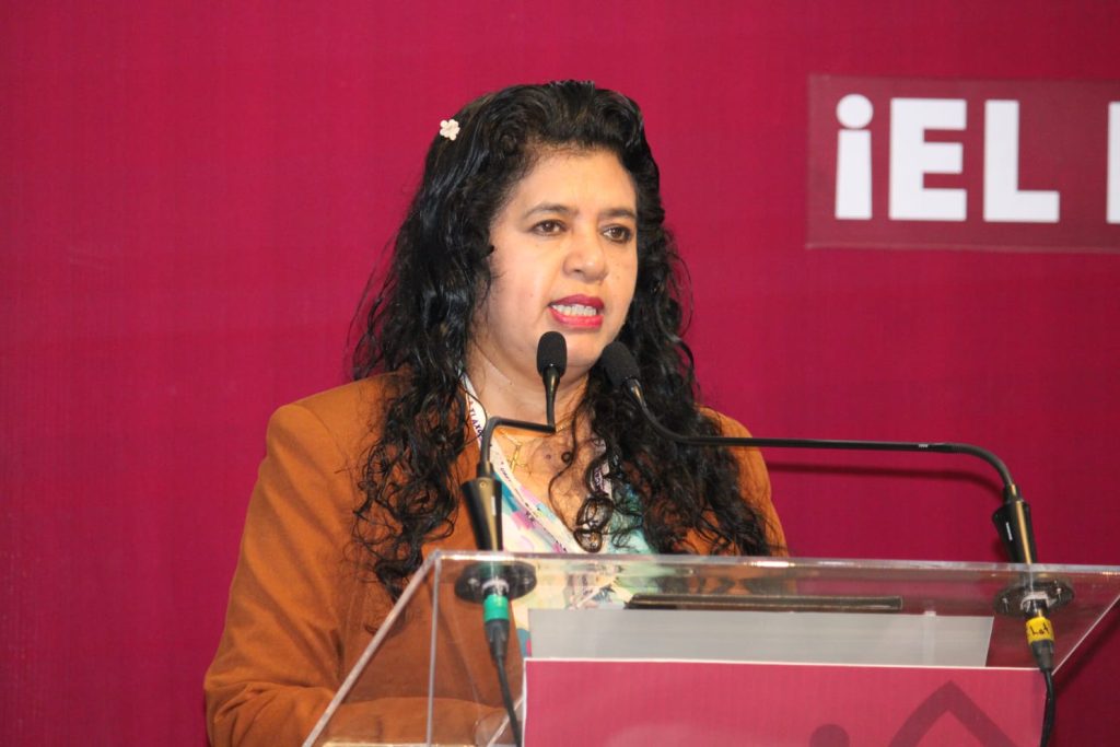 Mujer con cabello rizado y oscuro, vestida con blazer marrón, hablando en un micrófono durante una conferencia o evento en un fondo rosa con texto en español.