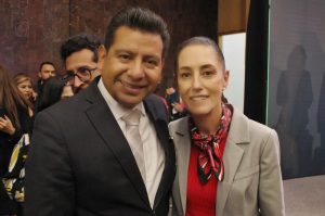Persona en un evento formal, vestido con traje y corbata, sonriendo para la cámara en un entorno con varias personas al fondo.