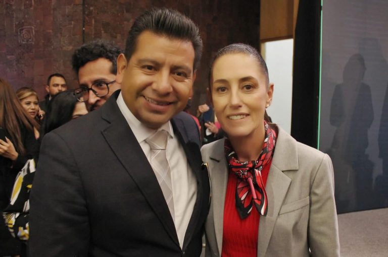 Persona en un evento formal, vestido con traje y corbata, sonriendo para la cámara en un entorno con varias personas al fondo.