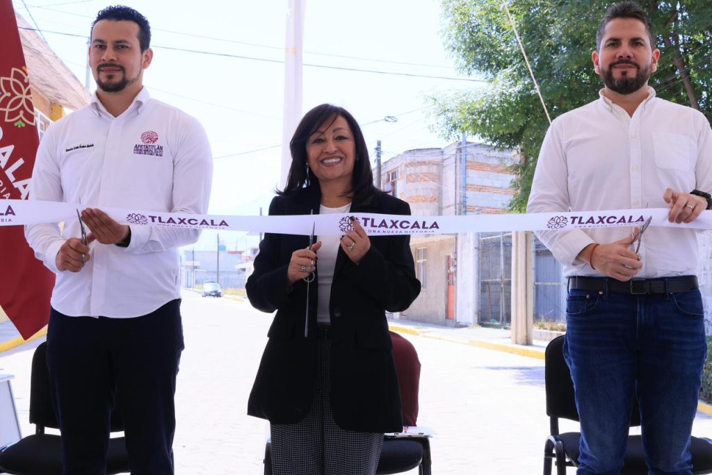 Corte de cinta en un evento oficial en Tlaxcala con tres personas sonriendo y sosteniendo la cinta, en un entorno urbano con árboles y edificios al fondo.
