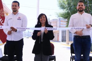 Corte de cinta en un evento oficial en Tlaxcala con tres personas sonriendo y sosteniendo la cinta, en un entorno urbano con árboles y edificios al fondo.