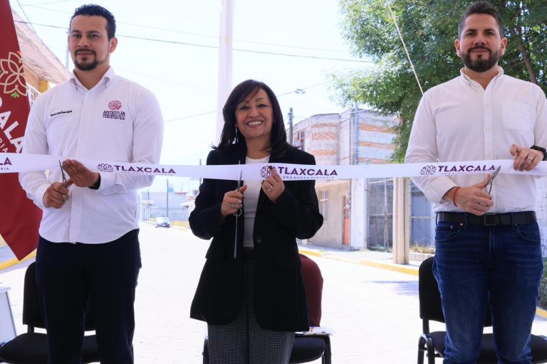 Corte de cinta en un evento oficial en Tlaxcala con tres personas sonriendo y sosteniendo la cinta, en un entorno urbano con árboles y edificios al fondo.