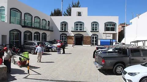 Edificio blanco de dos pisos con ventanas arqueadas y un pequeño balcón en la parte superior, en un área de estacionamiento con varias personas y vehículos.