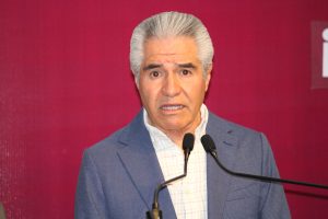 Hombre mayor con cabello gris, vestido con saco azul y camisa blanca, hablando en un micrófono en un evento formal.