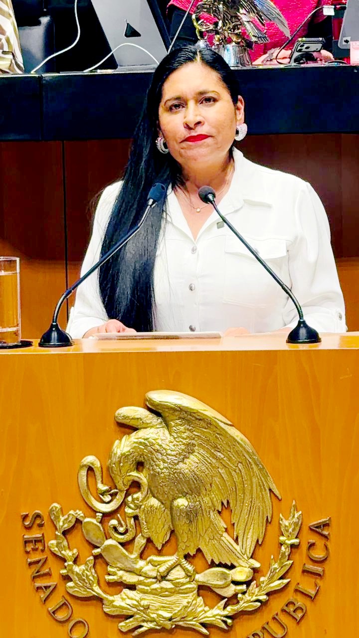 Mujer en un podio con escudo de Tlaxcala, hablando en un evento oficial, vestida con blusa blanca y cabello largo y oscuro.