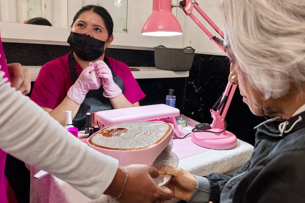 Manicurista atendiendo a una clienta en un salón de belleza, con herramientas y esmaltes en la mesa, en un ambiente bien iluminado y profesional.