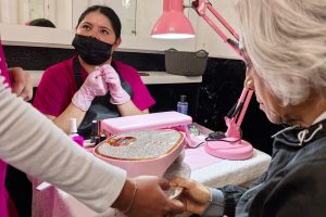 Manicurista atendiendo a una clienta en un salón de belleza, con herramientas y esmaltes en la mesa, en un ambiente bien iluminado y profesional.