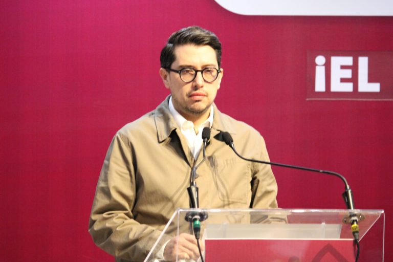 Hombre con gafas y chaqueta beige hablando en un podio durante un evento con fondo rojo y logotipo de iEL.