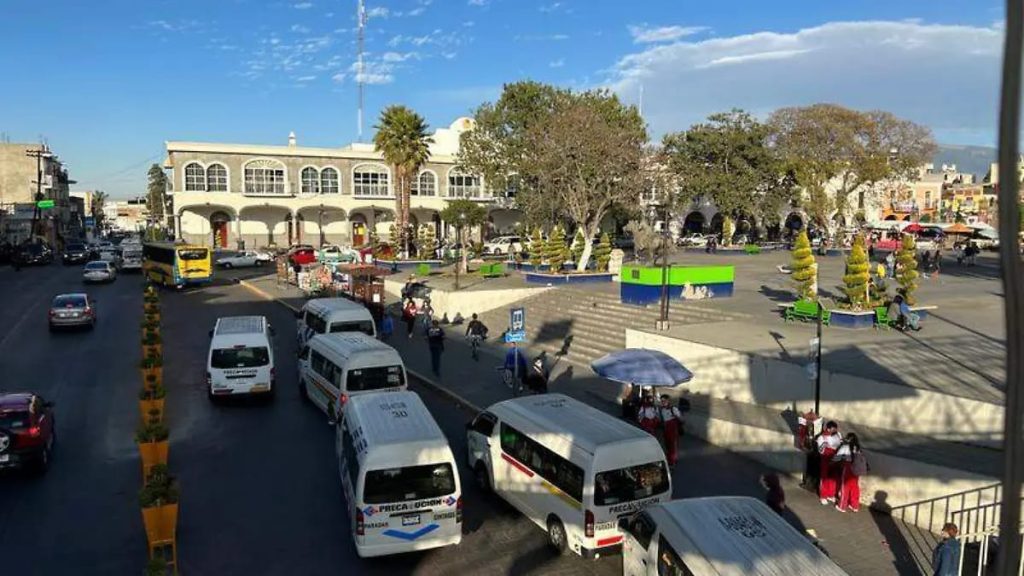 Plaza principal de Tlaxcala con vehículos y personas en un día soleado, rodeada de árboles y edificios históricos.