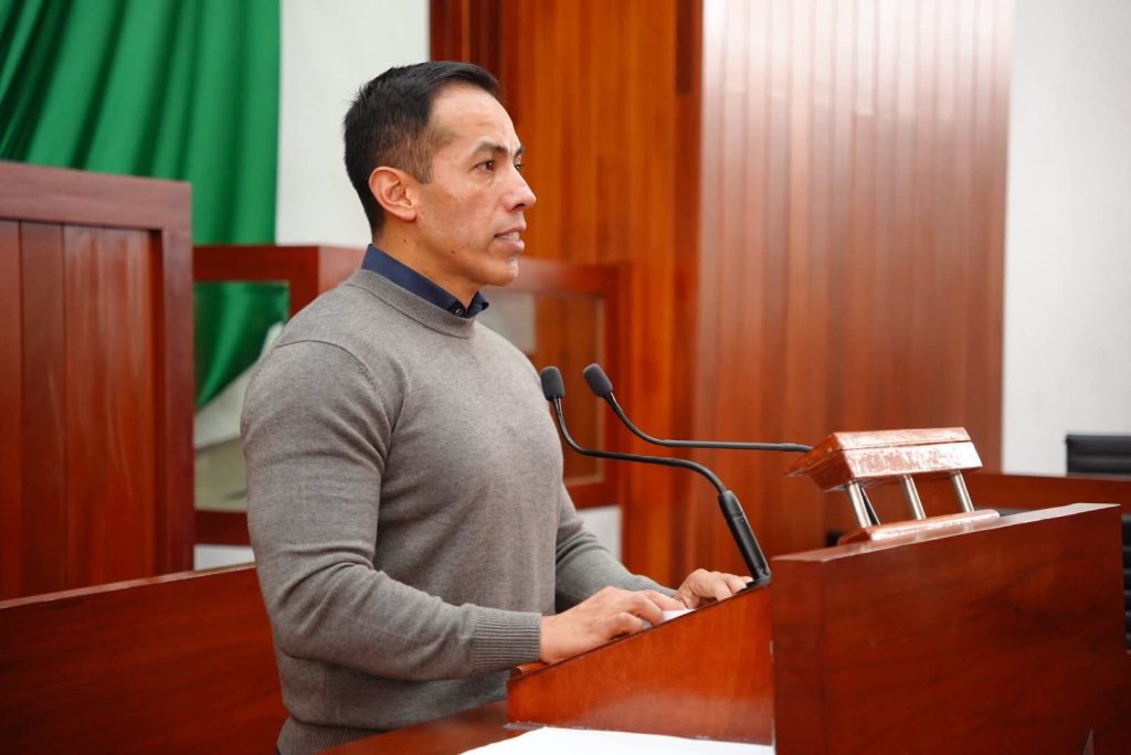 Hombre hablando en un podio en un entorno formal, con fondo de cortinas verdes y madera, durante una conferencia o evento oficial.