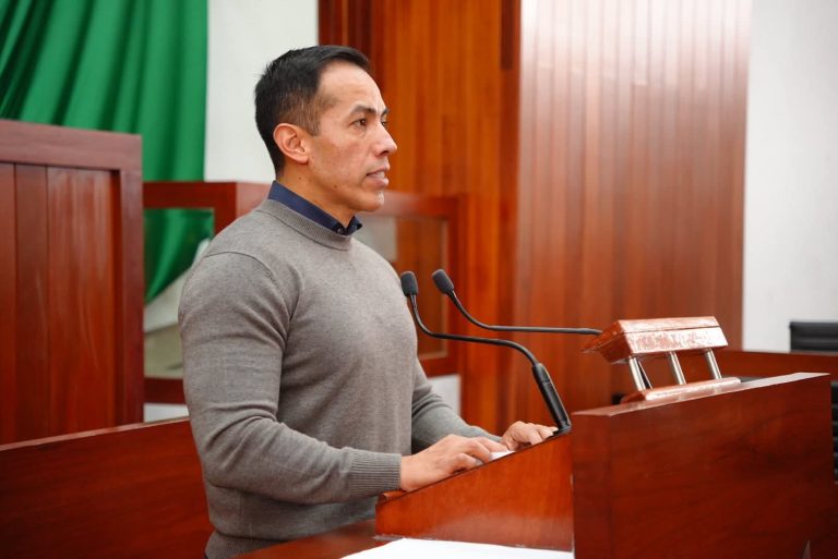 Hombre hablando en un podio en un entorno formal, con fondo de cortinas verdes y madera, durante una conferencia o evento oficial.