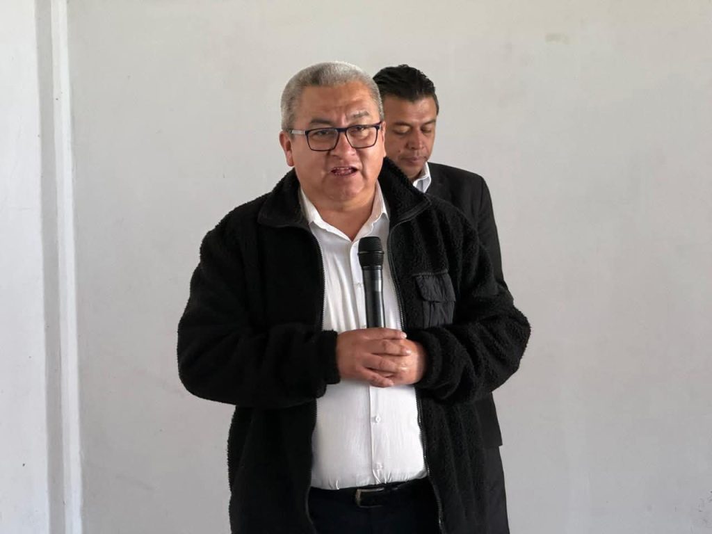 Hombre con gafas y cabello gris, sosteniendo un micrófono, en una conferencia o evento formal, con fondo blanco y otra persona en segundo plano.