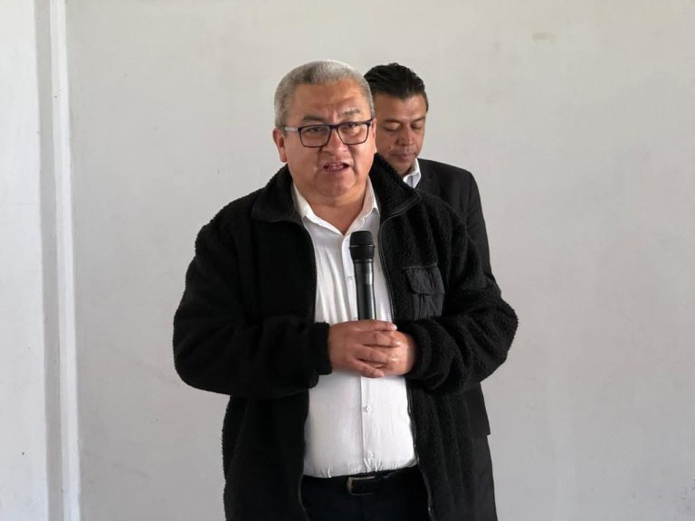 Hombre con gafas y cabello gris, sosteniendo un micrófono, en una conferencia o evento formal, con fondo blanco y otra persona en segundo plano.
