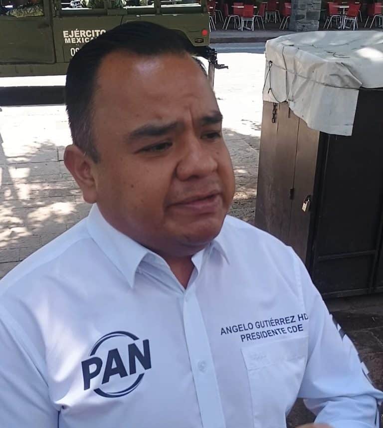 Hombre con peinado peinado hacia atrás, vestido con camisa blanca con logotipo del PAN y texto de su cargo, en un entorno al aire libre con sombra y mobiliario.