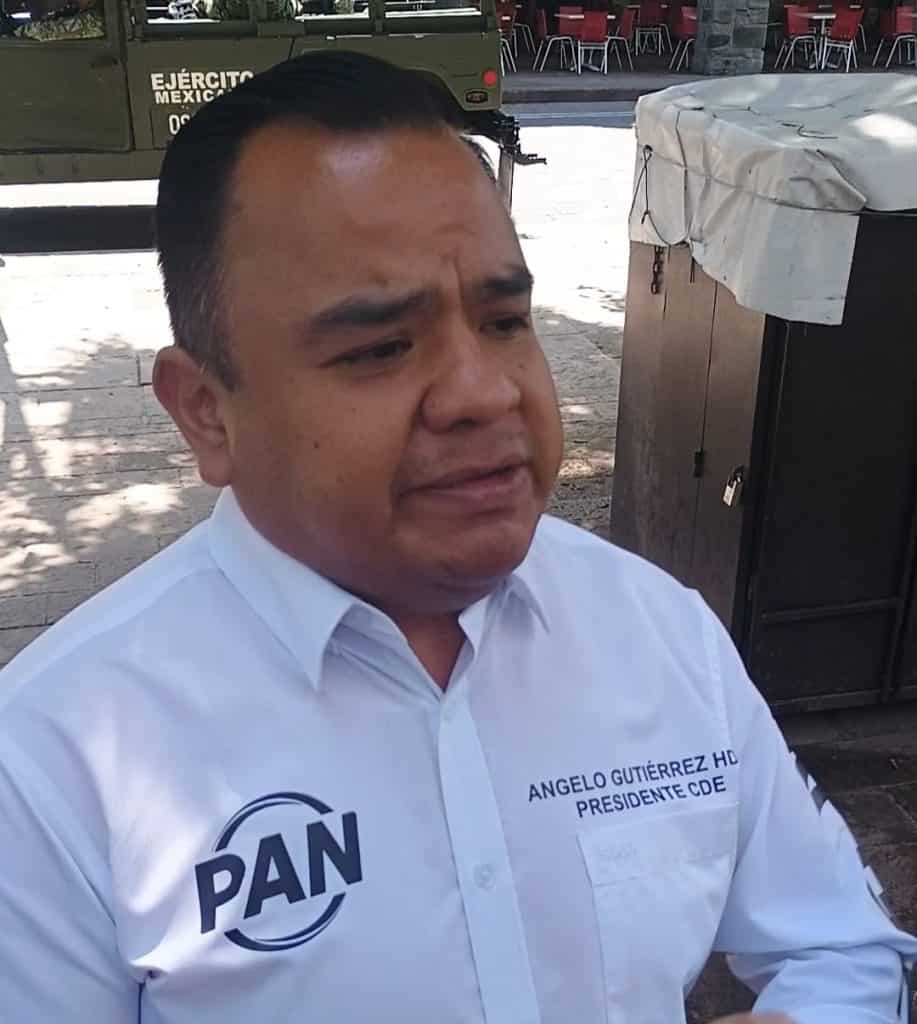 Hombre con peinado peinado hacia atrás, vestido con camisa blanca con logotipo del PAN y texto de su cargo, en un entorno al aire libre con sombra y mobiliario.