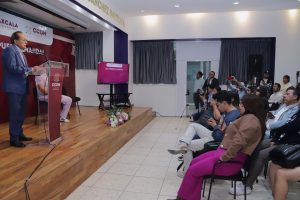 Hombre en traje dando discurso en conferencia frente a audiencia en sala de eventos con pantalla y micrófono.