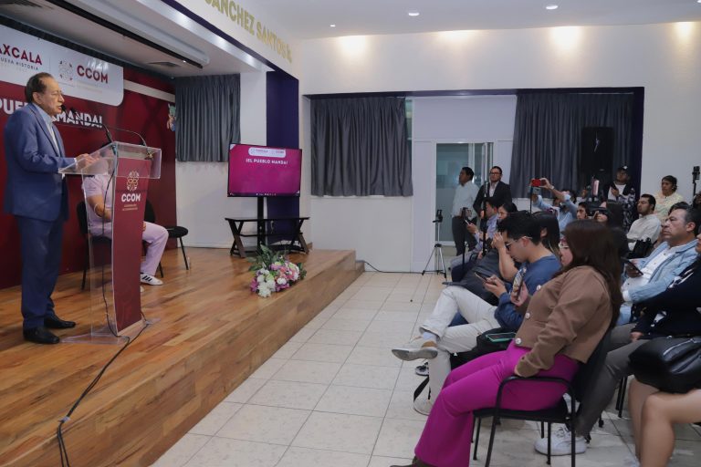 Hombre en traje dando discurso en conferencia frente a audiencia en sala de eventos con pantalla y micrófono.
