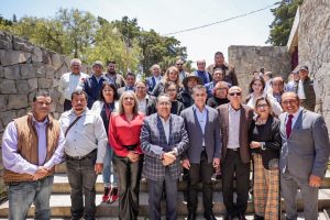 Persona grupo de adultos posando en un espacio al aire libre, algunos con gafas y ropa formal, en un entorno con muros de piedra y árboles al fondo.