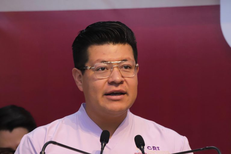 Árbol de etiquetas (ALT): Hombre hablando en un evento con micrófono, vestido con camisa blanca y gafas, en un fondo de color rojo y azul.