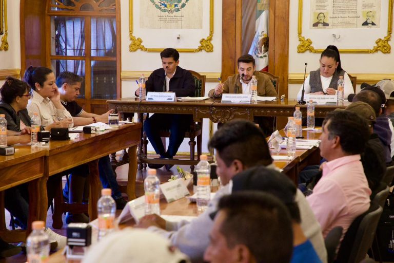 La reunión formal en una sala con paredes decoradas y cuadros, donde varias personas participan en una discusión o presentación oficial.