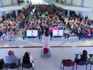Amplia audiencia en un evento público en Tlaxcala, donde una oradora presenta frente a un gran grupo de asistentes en un espacio cerrado con múltiples puestos y pantallas digitales.