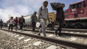 Hombres cruzando las vías del tren con equipaje y objetos personales en un entorno de estación ferroviaria. Personas en movimiento, algunos con mascarillas, en un día nublado en Tlaxcala, México.