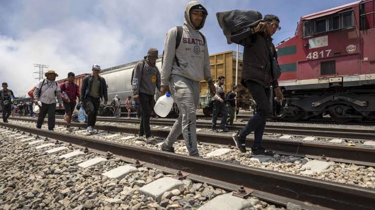 Hombres cruzando las vías del tren con equipaje y objetos personales en un entorno de estación ferroviaria. Personas en movimiento, algunos con mascarillas, en un día nublado en Tlaxcala, México.