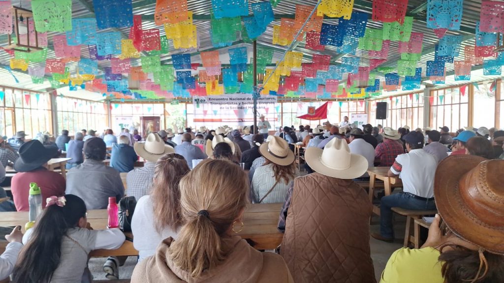 Cortometraje en un evento cultural con decoraciones coloridas y muchas personas asistiendo, incluyendo adultos y niños, en un espacio cubierto con ambiente festivo y comunitario.