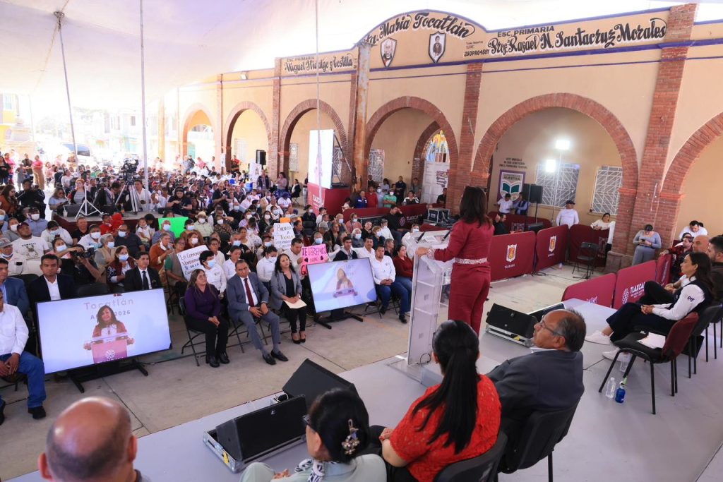 Conferencia o evento público con audiencia numerosa en un espacio cubierto con arcos de ladrillo y pantallas digitales.